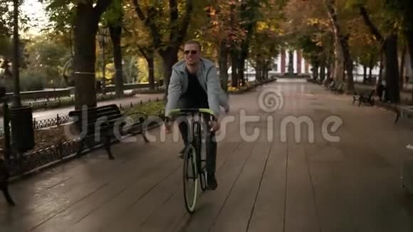 一个戴太阳镜的年轻微笑男子在早晨公园或林荫大道骑自行车的正面镜头慢动作视频的预览图