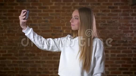 漂亮的白种人金发女人正在手机的网络摄像头上拍照站在砖头上放松的轮廓视频的预览图