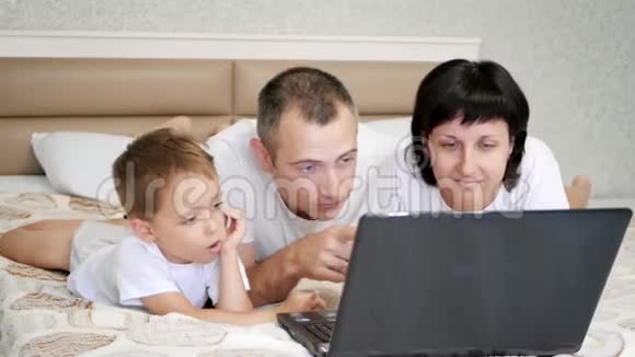幸福的一家人爸爸妈妈和孩子正躺在床上看着笔记本电脑微笑着爸爸亲吻他的妻子和儿子视频的预览图