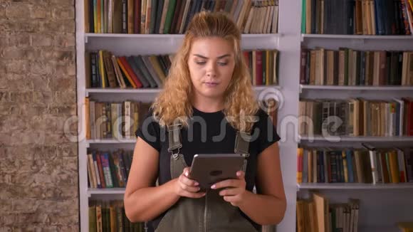 一位漂亮的白种人金发女性正在翻阅她的平板电脑而她则站在图书馆的书旁边专注于打字视频的预览图