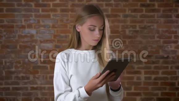 美丽的金发白种人女孩正对着镜头微笑在室内红砖工作室冰冷而有趣地擦拭着平板电脑视频的预览图