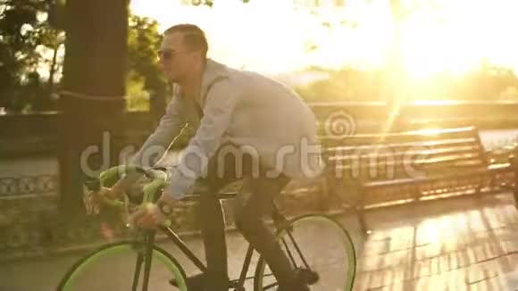 一位戴太阳镜的年轻微笑男子在早晨公园或林荫大道骑自行车的侧面景色年轻人的慢动作视频的预览图