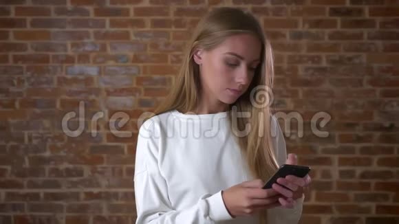 漂亮的金发白种人女孩在她的智能手机滚动精确地看同时站在砖墙旁边视频的预览图