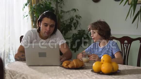 坐在厨房桌子上的小男孩和卡布奇诺一起吃牛角面包哥哥看着笔记本电脑视频的预览图