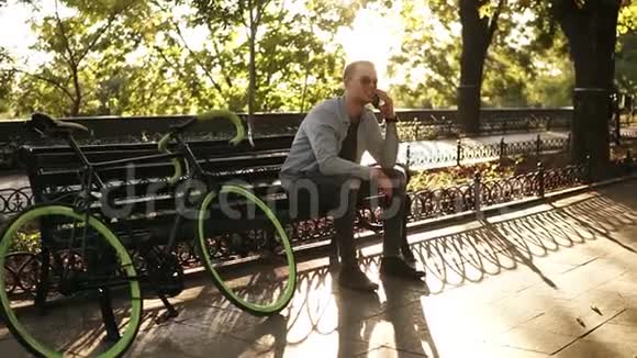 穿着蓝色衬衫的微笑男人坐在长凳上用手机说话骑自行车后休息视频的预览图
