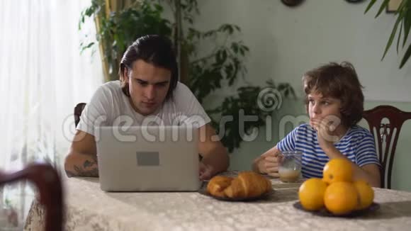 坐在厨房桌子上的小男孩和卡布奇诺一起吃牛角面包哥哥看着笔记本电脑视频的预览图