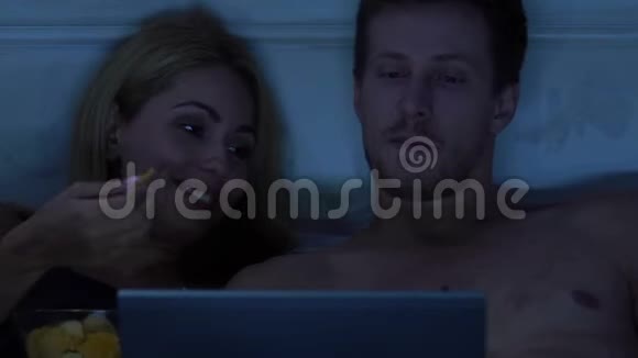 一对年轻夫妇在床上看着笔记本电脑上的连载吃零食喝啤酒视频的预览图