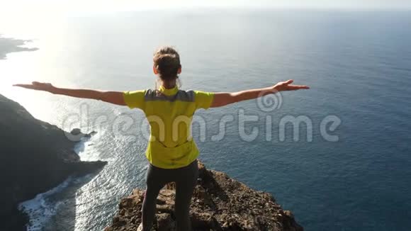 年轻女子举起双臂站在美丽的陡峭悬崖上在美丽的风景中登上顶峰的女士视频的预览图