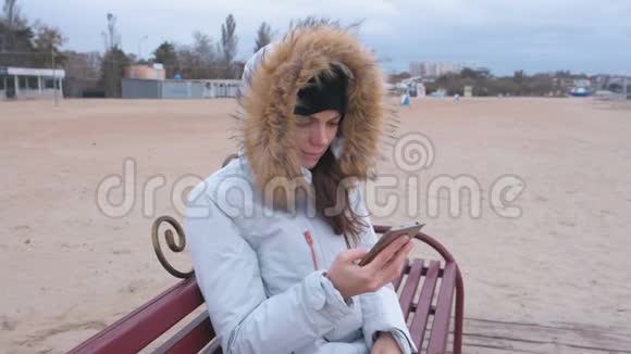 穿着白色羽绒服的女人坐在海滩上的长凳上看着手机上悲伤的视频视频的预览图