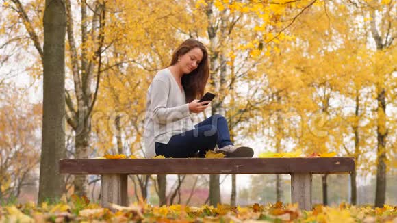 带着智能手机的女人在秋天公园的黄色枫树上放松视频的预览图
