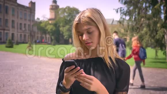 漂亮的白种人短发金发女性正在外面靠近老城区和公园的时候滚动手机视频的预览图