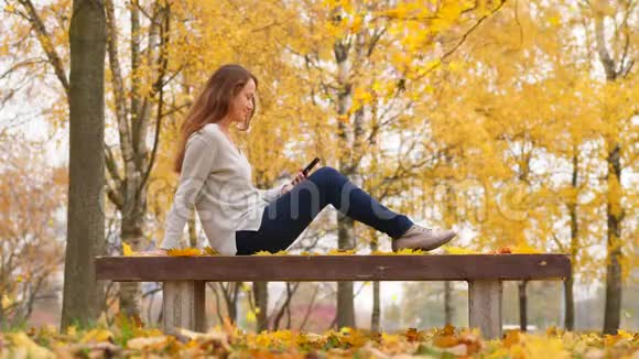 有智能手机的女人在秋天公园里躺在黄色的枫叶里休息视频的预览图