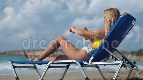 女孩躺在海滩上的休息室里用晒伤的喷雾在她的身上以免在阳光下被灼伤视频的预览图