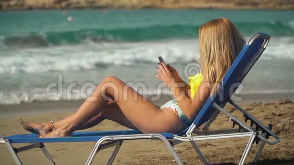 金发女人在海滩上晒日光浴躺在甲板椅上大海在后台女孩在手机上发短信休闲视频的预览图
