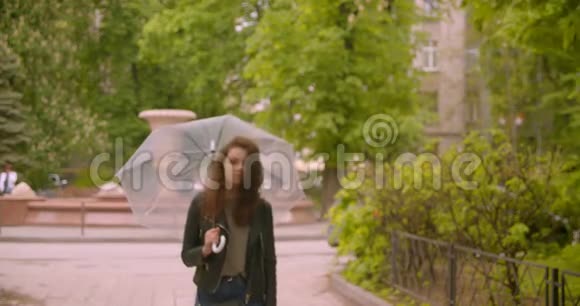 在绿色公园里白种人深色头发的女性带着雨伞微笑着走进镜头视频的预览图