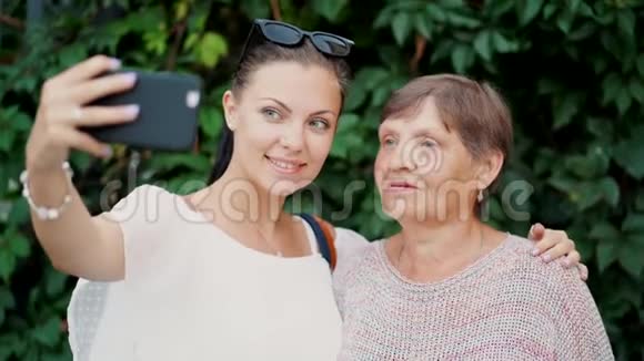 美丽的老奶奶和她的孙女在树叶背景下户外自拍看着相机微笑着视频的预览图