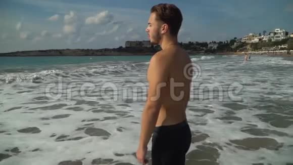 阳光明媚的一天站在风景如画的美丽海滩上半裸的运动男子向海浪走去慢动作视频的预览图