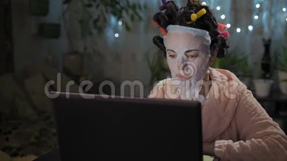 年轻漂亮的女士戴着恢复活力的面具和卷发器看着笔记本电脑屏幕视频的预览图