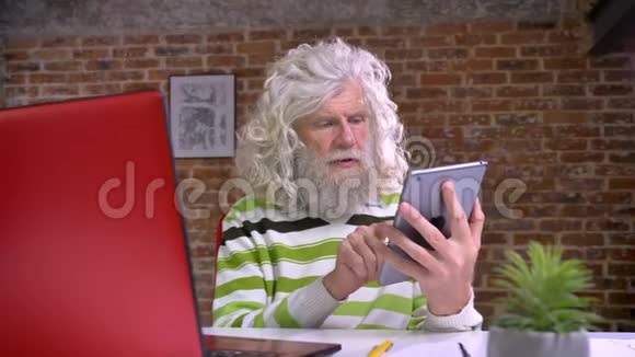 专注的白种人老奶奶长着浓密的胡须和长长的白发正在擦拭他的平板电脑集中注意力看着视频的预览图