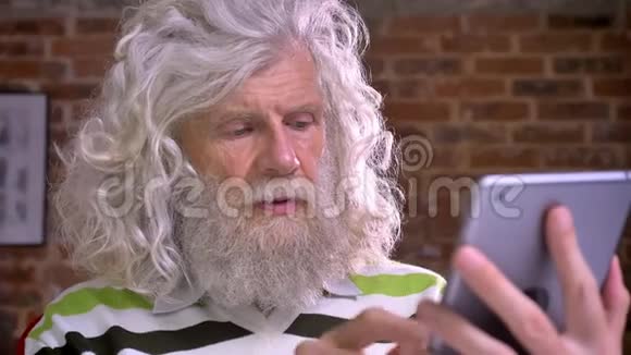 古代白种人长着令人敬畏的白胡子长着一头浓密的头发在现代砖房的平板上打字视频的预览图