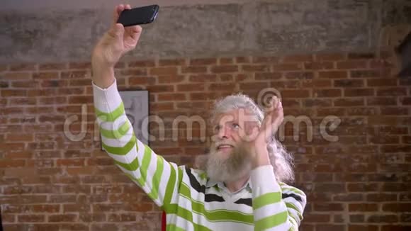 可爱的老白种人男性正在向智能手机的前照相机挥手微笑着放松长长的白发砖头视频的预览图
