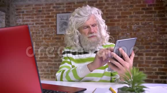 快乐的老白种人留着白大胡子和波浪发在他的平板电脑上展示着蓝色的屏幕视频的预览图