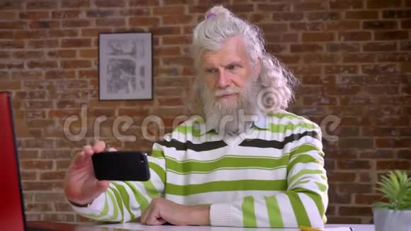 有趣的老白种人有着令人敬畏的白色长发和胡须坐在桌面上随意自拍视频的预览图
