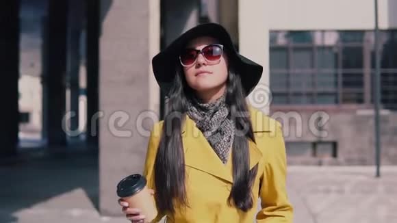穿着黄色雨衣帽和眼镜走在街上喝咖啡的时髦女孩视频的预览图