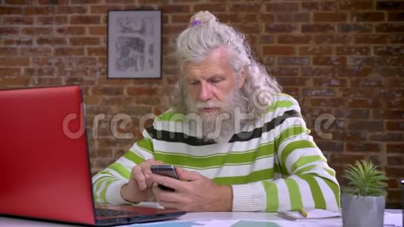 酷酷的白种人老人坐在工作场所一边滚动着手机身后是砖墙长长的白色视频的预览图