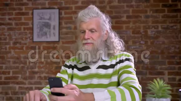 令人惊叹的现代祖父在红砖办公室里一边用网络摄像机一边说话一边拿着手机四处走动视频的预览图