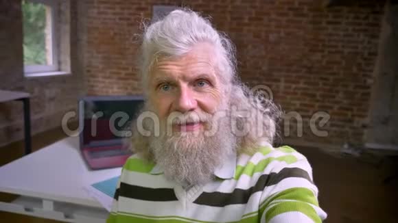 可爱的老男人留着白色的头发和长长的胡须拿着相机自拍同时站在砖头办公室制作视频的预览图