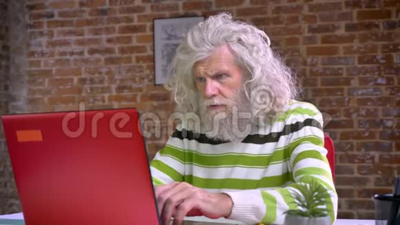 一位留着白长胡子和头发的老奶奶坐在砖房里专心致志地打着红电脑视频的预览图