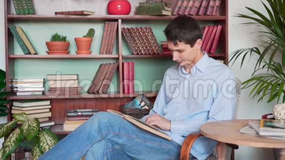 一个年轻人坐在家里的图书馆里看书一个穿着蓝色衬衫和牛仔裤的人拿着一本书看书视频的预览图