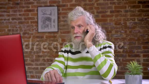 忙碌的可爱的长着胡子的白种人老人挂在电话上一边看着电脑一边说着冷话视频的预览图