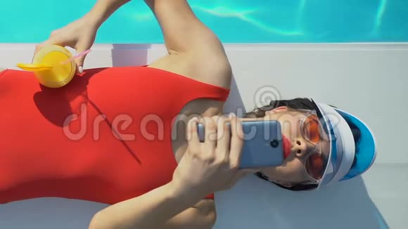 时尚女性在泳池附近使用手机喝鸡尾酒在网上写博客视频的预览图