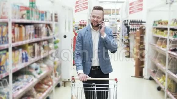 穿着白色衬衫的年轻微笑男子正在智能手机上交谈并在超市的货架之间行走着商品视频的预览图