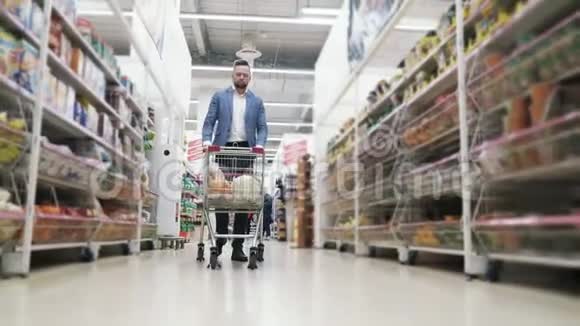 男人卷手推车与食品在货架和选择产品在超市稳定射击视频的预览图