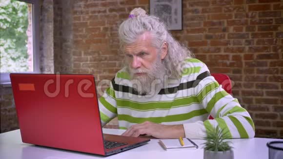 可爱的白种人老人坐在他的电脑旁边写着笔记白发和胡须随意而寒冷视频的预览图