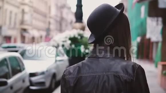 镜头跟随年轻的女性时尚博主穿着旧的皮夹克和时尚的帽子沿着城市街道慢动作视频的预览图