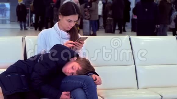 女人带着她疲惫的儿子在机场大厅手里拿着手机等待航班视频的预览图