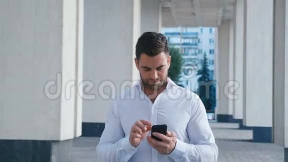 年轻有魅力的商人走在办公楼附近在他的智能手机上输入一条信息看上去很成功视频的预览图