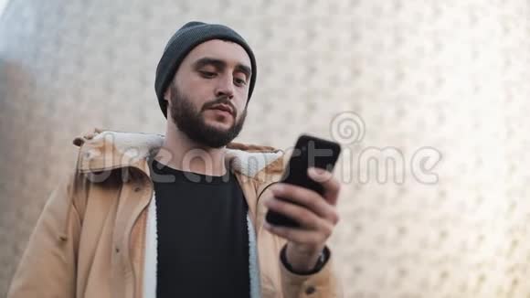 年轻的胡须男在户外用手机发送音频信息他穿着一件秋装和一顶针织帽子视频的预览图