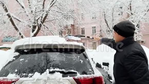 一名年轻人用伸缩刷子从暴风雪后的雪中清洁汽车的4K镜头视频的预览图