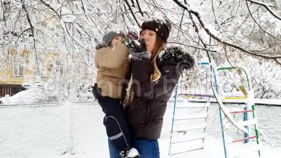 4k视频快乐微笑的年轻女子抱着她蹒跚学步的男孩在冬季公园玩雪视频的预览图
