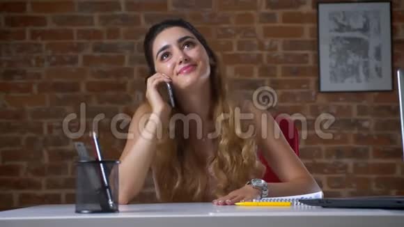 可爱的白种人女孩在办公室里坐在靠近笔记本电脑的桌面上轻松地用手机说话工作轻松视频的预览图