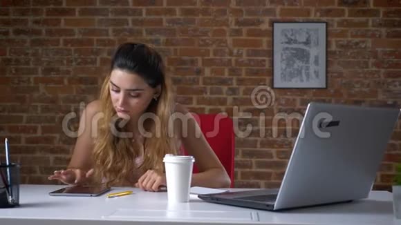 漂亮的白种人女性正在擦拭她的平板电脑喝着白色杯子里的咖啡被隔离在砖工作室里视频的预览图