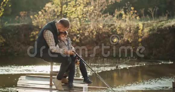 年轻的爸爸和他的儿子在湖边钓鱼他们有一个快乐的心情一起微笑度过一段可爱的时光视频的预览图
