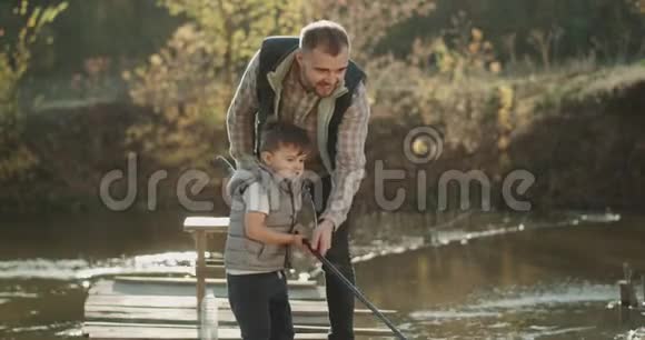 肖像爸爸鼓励他的儿子在阳光明媚的日子在湖边钓鱼一起度过美好的时光视频的预览图
