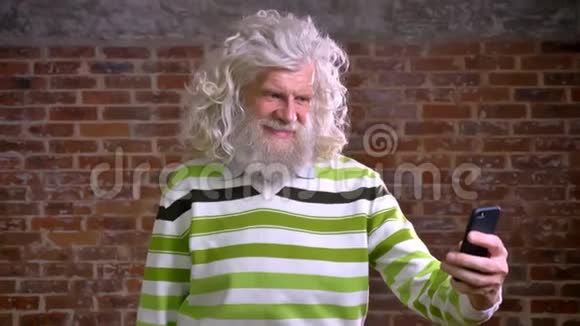 快乐的退休白种人男性留着白厚胡子和长发站在阉割的附近打着视频电话视频的预览图