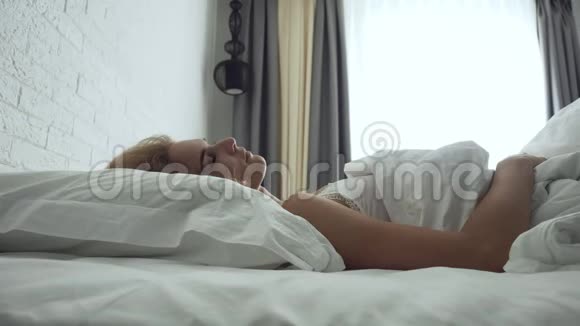 年轻女子醒来在舒适舒适的床上柔软的白色枕头和床单视频的预览图